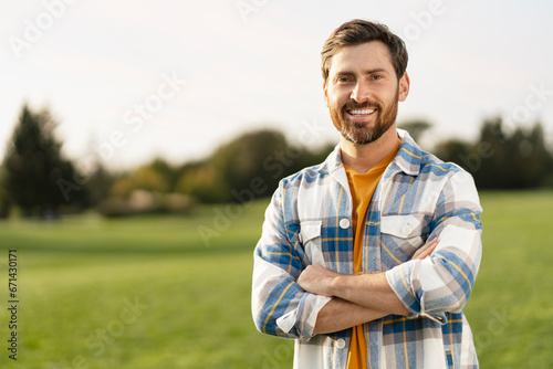 Portrait of smiling handsome middle aged man farmer wearing s stylish t shirt holding arms crossed looking at camera standing in green field, copy space. Successful business concept