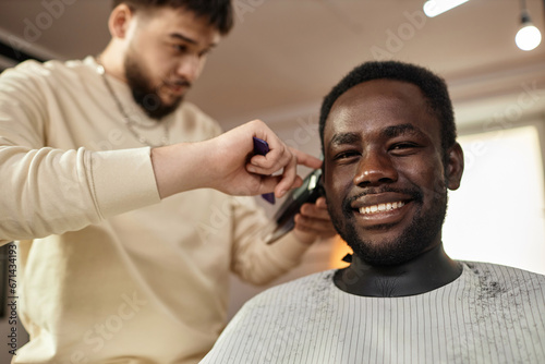 Portrait of African American man smiling at camera while barber cutting his hair with trimmer and comb