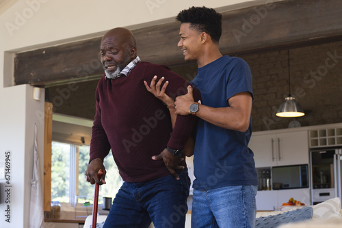 Happy african american adult son helping senior father stand up from couch at home photo