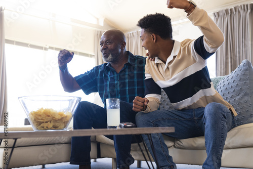 Excited african american father and adult son watching sport on tv and celebrating victory photo