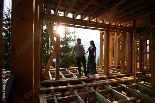 Man and woman inspecting their future wooden frame house nestled in the mountains near forest. Youthful couple at construction site in early morning. Concept of contemporary ecological construction.