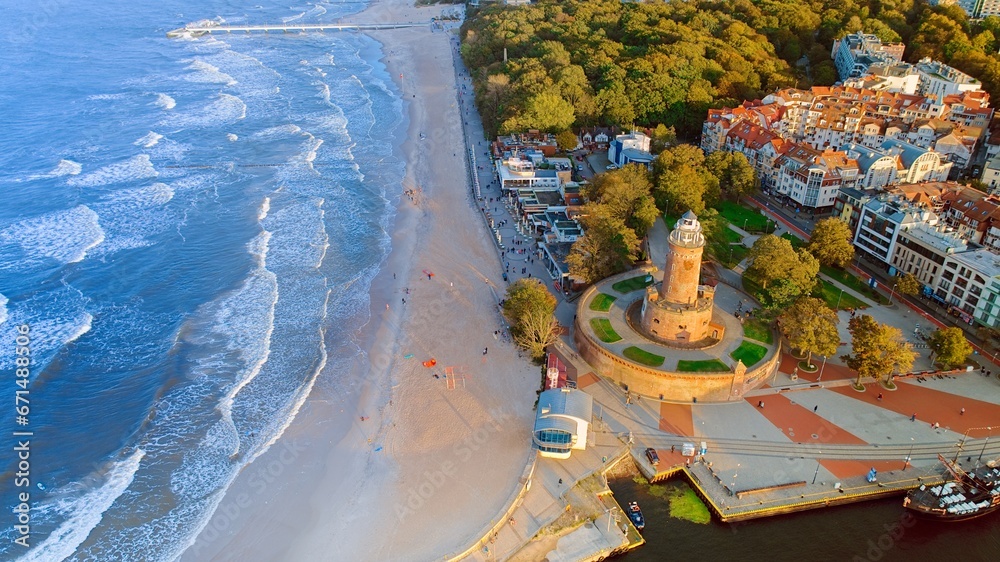 Drone photo captures Kołobrzeg's maritime charm, featuring the iconic lighthouse, cerulean sea, turbulent waves, a distant pier, and autumnal hues on the trees. - obrazy, fototapety, plakaty 