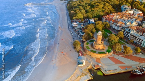 Drone photo captures Kołobrzeg's maritime charm, featuring the iconic lighthouse, cerulean sea, turbulent waves, a distant pier, and autumnal hues on the trees. photo