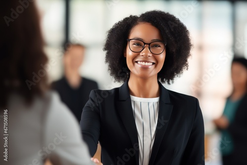Young African American businesswoman in eyeglasses with kinky hair smiles greeting colleagues. Cheerful business lady meets new employees. Joyful young woman in formal wear shakes hand of co-worker.