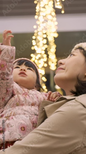 Vertical Screen: slow motion of cute curious asian toddler baby girl raising up hand trying to touch outdoor Christmas lights in her motherâs arms outside an urban shopping mall