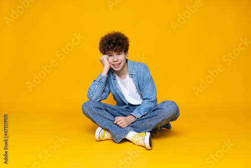Curly boy teenager sitting on the floor, on a yellow background. © Atlas