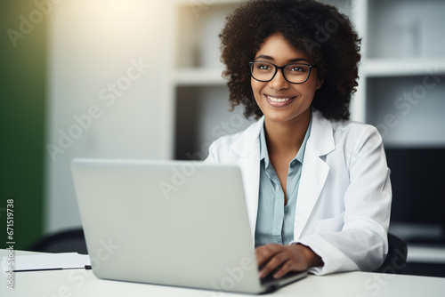 African American woman doctor sitting at desk and working on laptop computer in modern bright hospital ordination. Smiling at camera.