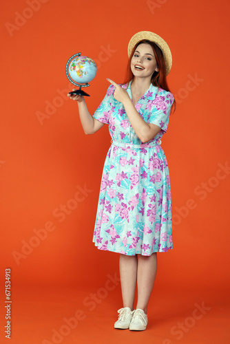 Woman in hat and dress looking at the camera smiling, holding small globe and pointing at it, isolated on orange background. Portrait of female traveler. © kinomaster