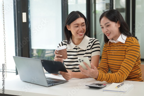 Two diverse mixed race beautiful african and asian business women wearing casual clothes, smiling with happiness, talking while taking break in indoor office, looking at laptop, holding cup of coffee