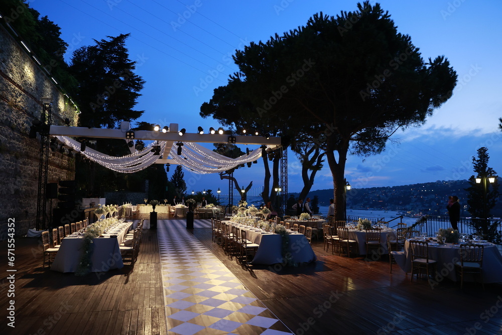 Decorated white wedding table for a festive dinner with pink flowers in brass pots on green lawn under the open sky.