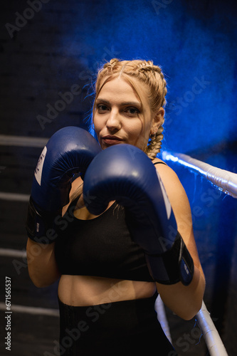 Strong female fighter in blue boxing gloves is training in the ring against a blue light background