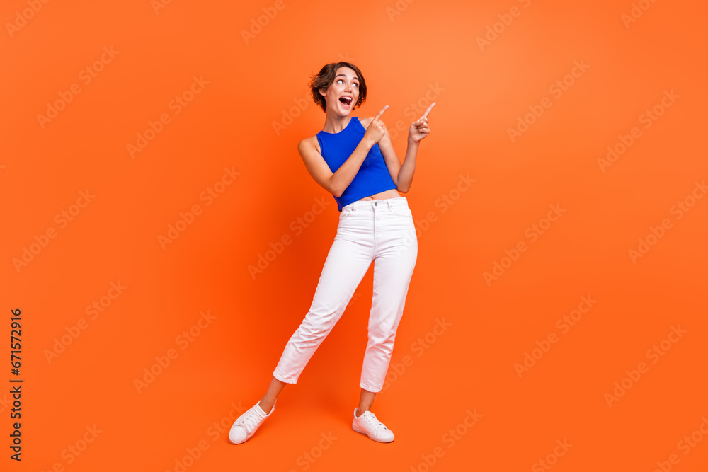 Full size portrait of astonished pretty girl look direct fingers empty space information isolated on orange color background Full size portrait of astonished pretty girl look direct fingers empty space information isolated on orange color background