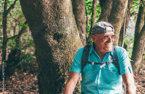 Portrait of smiling senior man with sunglasses and backpack sitting in a trekking day in the forest enjoying nature and healthy lifestyle in vacation or retirement