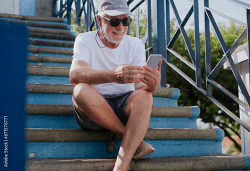 Smiling handsome bearded man in hat and sunglasses while using phone sitting on outdoor staircase. Elderly male enjoying free time, tech and social #671576771