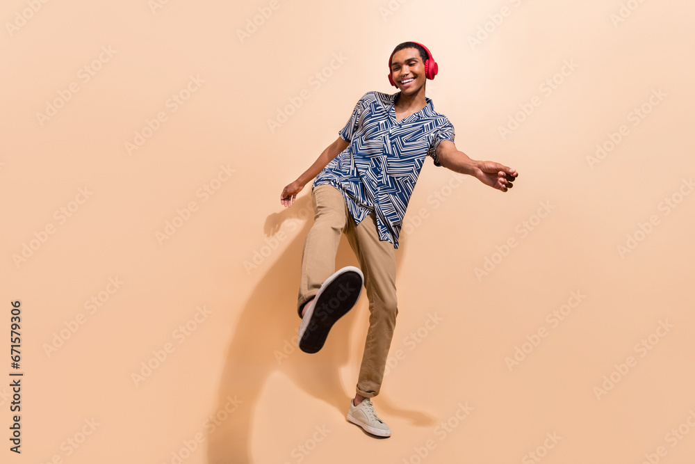 Full length photo of positive cheerful man dressed stylish shirt brown pants in earphones dancing isolated on beige color background