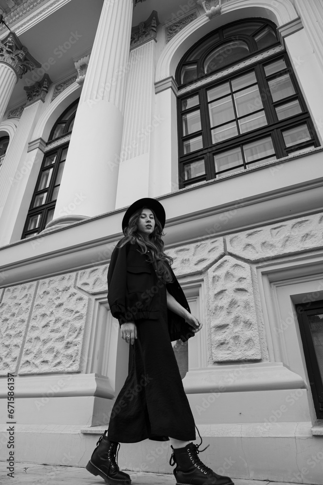 Black and white image of a young beautiful stylish woman walking in black clothes, posing near museum.