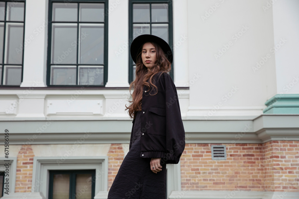 Attractive young beautiful fashionable woman posing in black hat and jacket, outside.