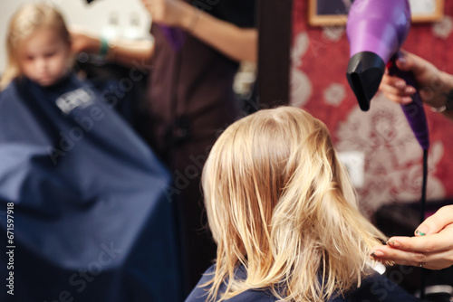 Hair salon, hairdresser blow dry hair for kid in barber shop at mirror. Barber woman make fashionable hairstyle for cute little blonde girl in barbershop. Haircare, beauty concept. Copy ad text space