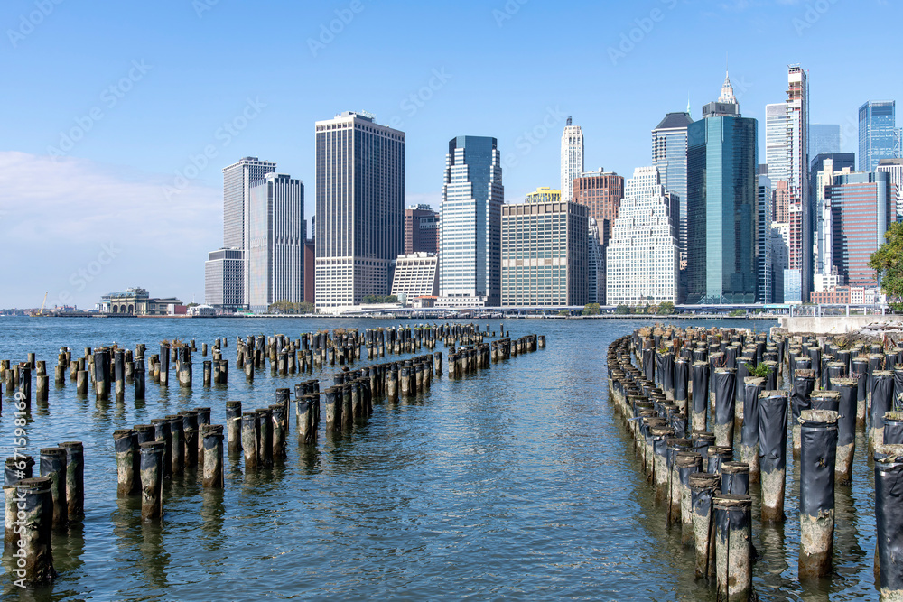Obraz premium Hundreds of old wooden pier supports in the East River with panoramic view on skyscrapers and landmarks of Lower Manhattan, New York City, NY, USA against a clear blue sky