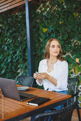 Young woman in casual clothes drinking coffee and talking on the phone, working on a green terrace at a laptop, looking at the screen and working on a project remotely from work in a cafe