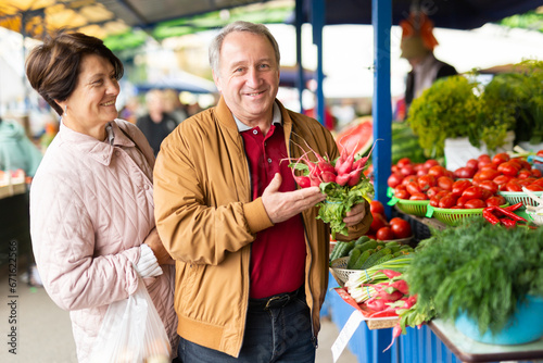 Positive mature couple in casual wear picking fresh radish during shopping at crowded bazaar photo
