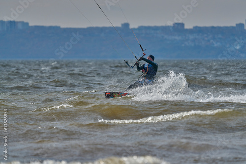  A mature male surfer rides a board with a kite on a windy autumn day across a large body of water. Splashes of water scatter to the sides.