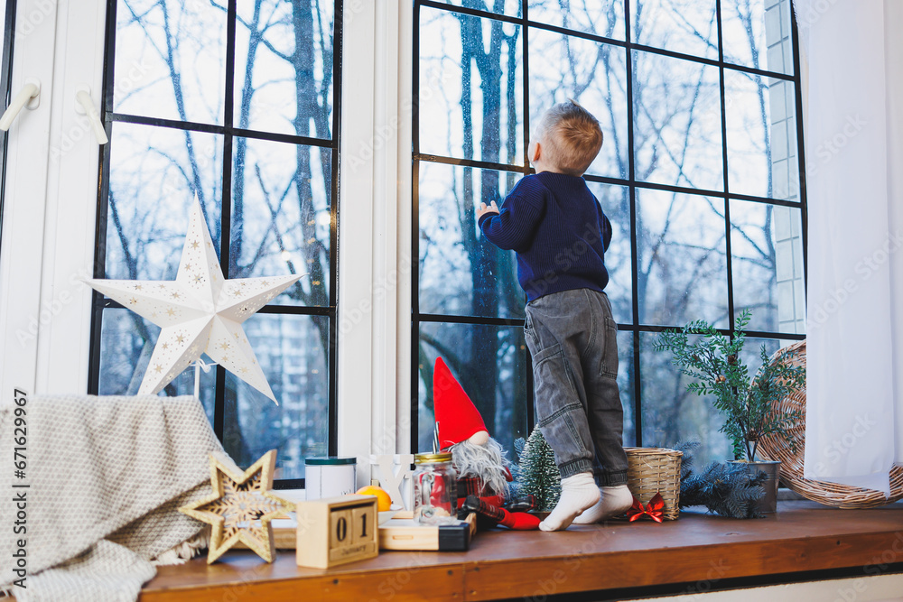 Fototapeta premium A 2-year-old boy sits on the windowsill in the new year. New Year's atmosphere at home. The child is waiting for the new year.