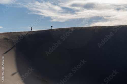 People hike up High Dune at Great Sand Dunes National Park in Colorado on a sunny summer evening, with mountains in the background