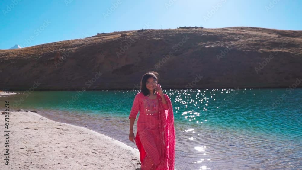 Portrait of attractive and young Indian woman smiling in Pink ethnic dress while walking against beautiful Chandra Taal lake in Spiti Valley, India. Beautiful young woman at turquoise color lake. Portrait of attractive and young Indian woman smiling in Pink ethnic dress while walking against beautiful Chandra Taal lake in Spiti Valley, India. Beautiful young woman at turquoise color lake.