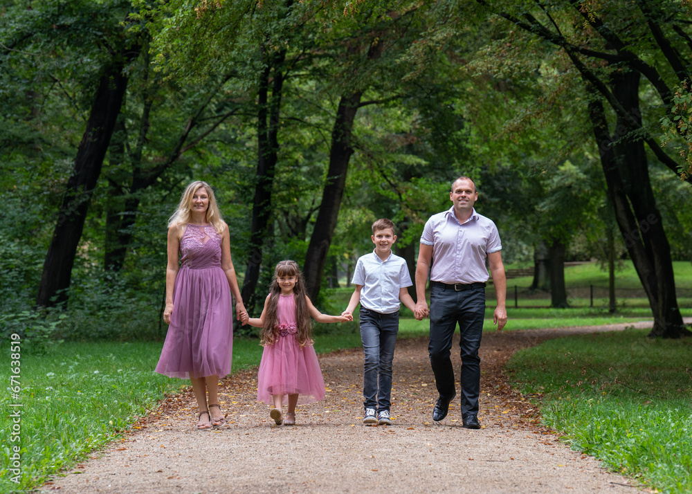 Portrait of a happy family. Laughing dad and mom, teenage son and first-grader daughter hold hands and walk along a path in the park. Mom and girl are dressed in beautiful pink dresses. Portrait of a happy family. Laughing dad and mom, teenage son and first-grader daughter hold hands and walk along a path in the park. Mom and girl are dressed in beautiful pink dresses.