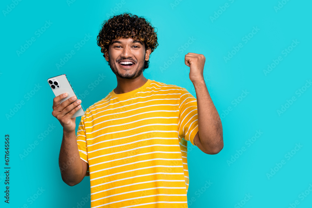 Photo portrait of nice young male fist device celebrate sales wear trendy striped yellow garment isolated on aquamarine color background Photo portrait of nice young male fist device celebrate sales wear trendy striped yellow garment isolated on aquamarine color background