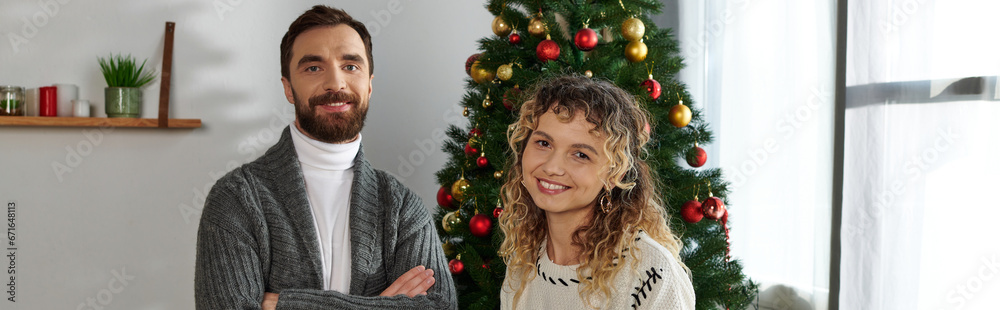 happy couple in winter clothing standing near decorated Christmas tree in modern apartment, banner