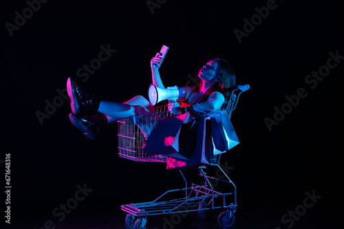 Attractive woman sitting in shopping cart holding smartphone and megaphone, smiling taking selfie. Isolated on black background in neon light.