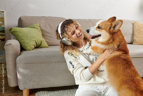 curly woman in wireless headphones playing with cute corgi dog in modern apartment, happy moments photo