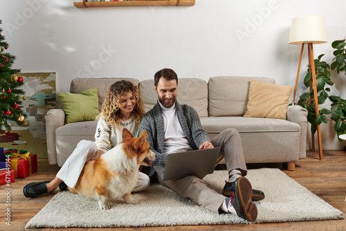 bearded man using laptop and sitting on carpet with curly wife near corgi dog and Christmas tree © LIGHTFIELD STUDIOS