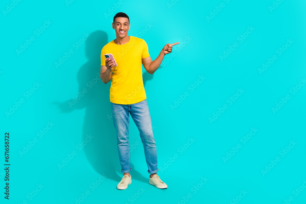 Full length portrait of cheerful handsome guy hold smart phone direct finger empty space isolated on turquoise color background Full length portrait of cheerful handsome guy hold smart phone direct finger empty space isolated on turquoise color background