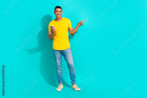 Full length portrait of cheerful handsome guy hold smart phone direct finger empty space isolated on turquoise color background