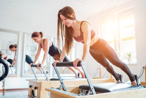 Two young females doing extended plank static strengthening core muscles exercise using pilates reformer machine in sport athletic gym hall. Active people training, yoga classes concept. photo