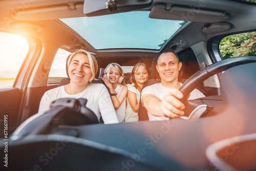 Happy young couple with two daughters inside car during auto trop. They are smiling, laughing during road trip. Family values, traveling concepts.