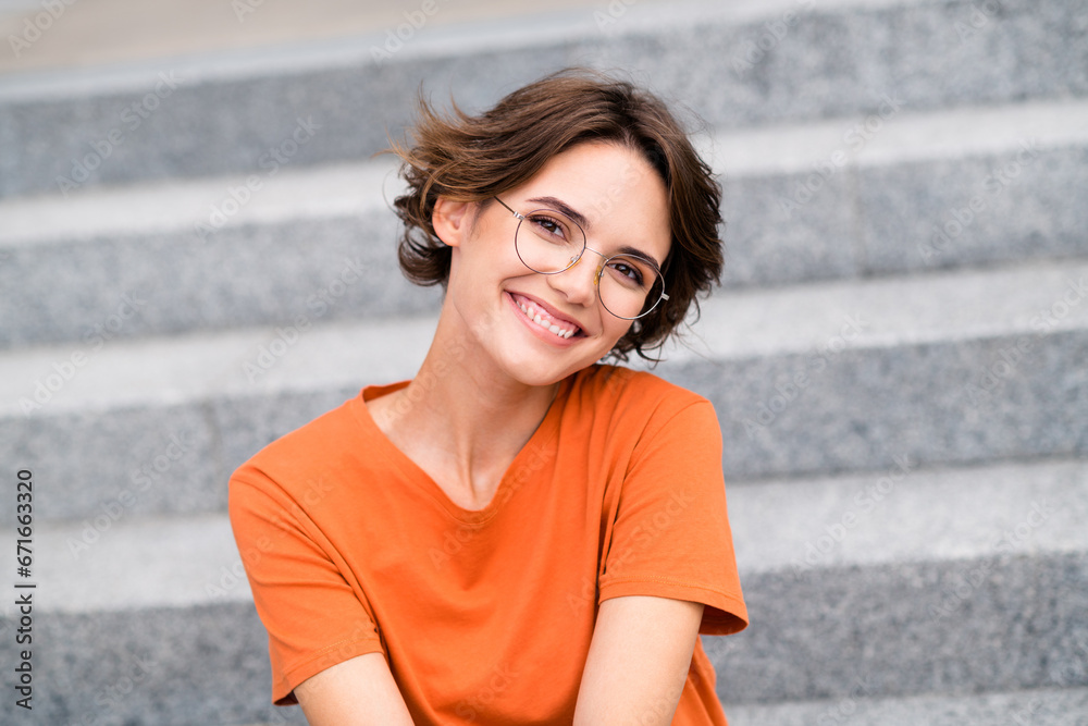 Photo of positive charming girl dressed orange t-shirt eyewear smiling sitting stairs outdoors urban town park Photo of positive charming girl dressed orange t-shirt eyewear smiling sitting stairs outdoors urban town park