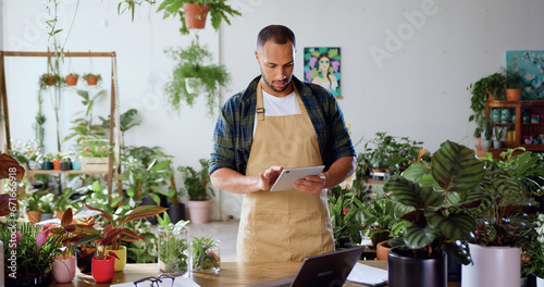 African American man checking green plants in flower shop and using tablet touching screen. Florist manager browsing on device in floral greenhouse. People and business concept.