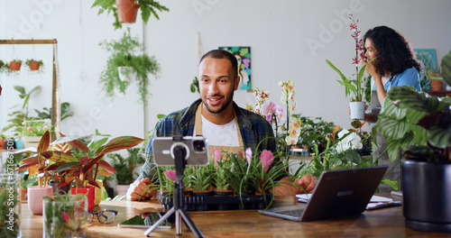 Handsome man gardener in apron using phone and recording blog video with clients. Young adult content creator influencer blogger recording a video talking about indoor plants care from workplace.
