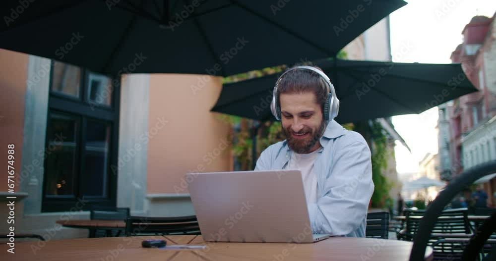 Stylish student working on laptop computer at college destination outdoors on terrace. Young man in headphones working at freelance during break in a cafe on sunny warm day. Online education concept. Stylish student working on laptop computer at college destination outdoors on terrace. Young man in headphones working at freelance during break in a cafe on sunny warm day. Online education concept.