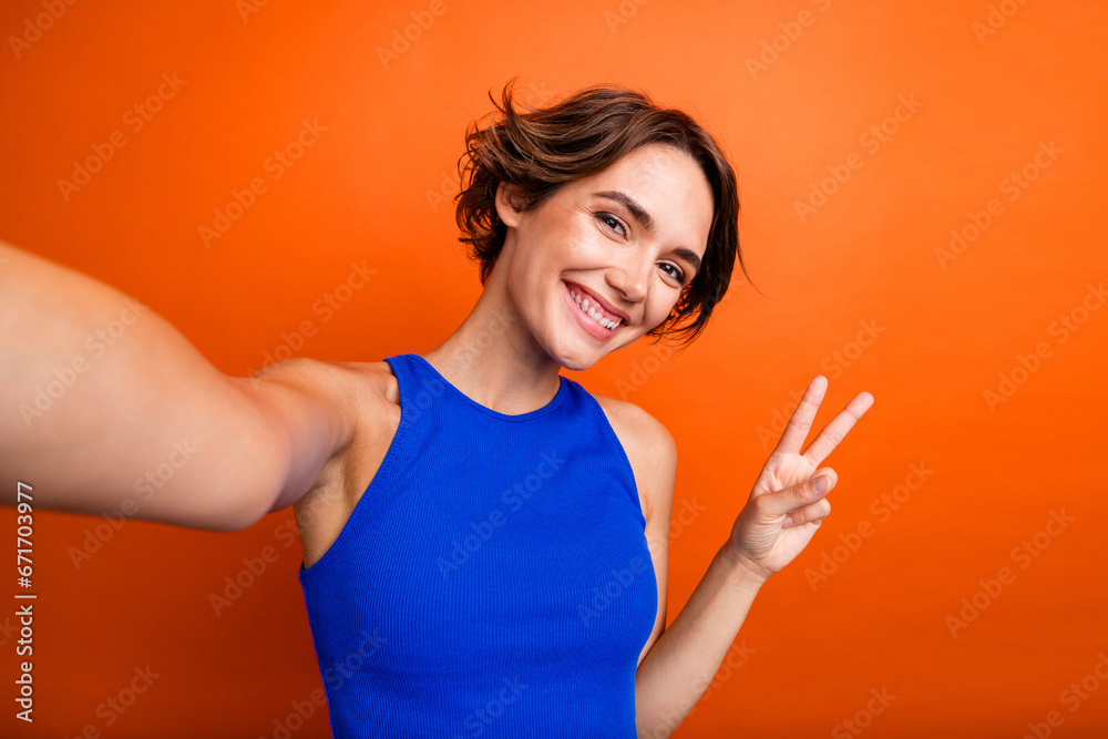 Photo of cheerful good mood lady wear blue singlet showing v-sign recording video vlog isolated orange color background Photo of cheerful good mood lady wear blue singlet showing v-sign recording video vlog isolated orange color background