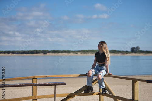 Young, pretty, blonde woman, wearing a black top, ripped jeans and tattoos, sitting on a railing, looking at infinity with the sea in the background. Concept of peace, tranquility, relaxation.