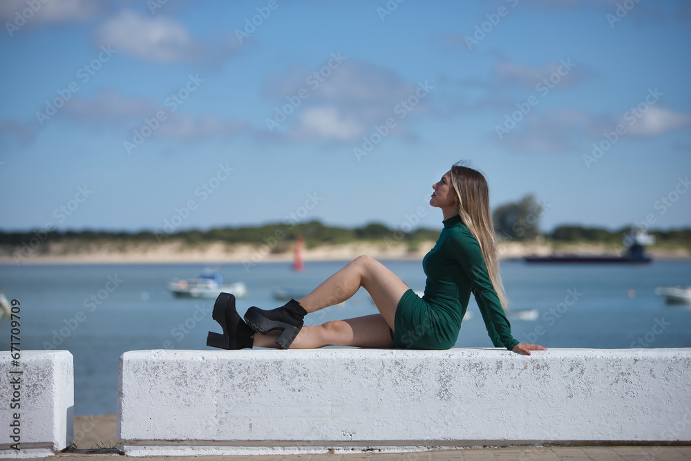 Young, beautiful, blonde woman, green eyes and wearing a green dress, sitting on a stone bench, solitary, relaxed and calm, with the sea in the background. Concept of solitude, peace, tranquility.