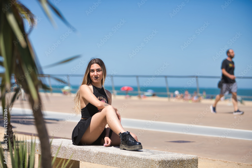 Young woman, beautiful and blonde, green eyes, with black top and skirt, tattoos, sitting on a stone bench, looking at the camera happy and relaxed. Concept looks, happiness, peace, relaxation.