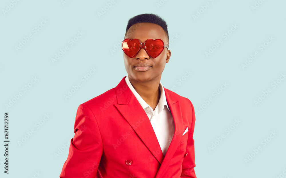 Portrait of handsome young African American man wearing stylish red suit and heart shaped sunglasses standing isolated on light blue background. Fashion and St Valentine's Day concept