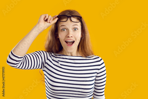 Portrait of funny positively amazed young woman looking at you with shocked expression. Caucasian woman raises glasses in surprise looking directly at you on orange background. Close up. Web banner.
