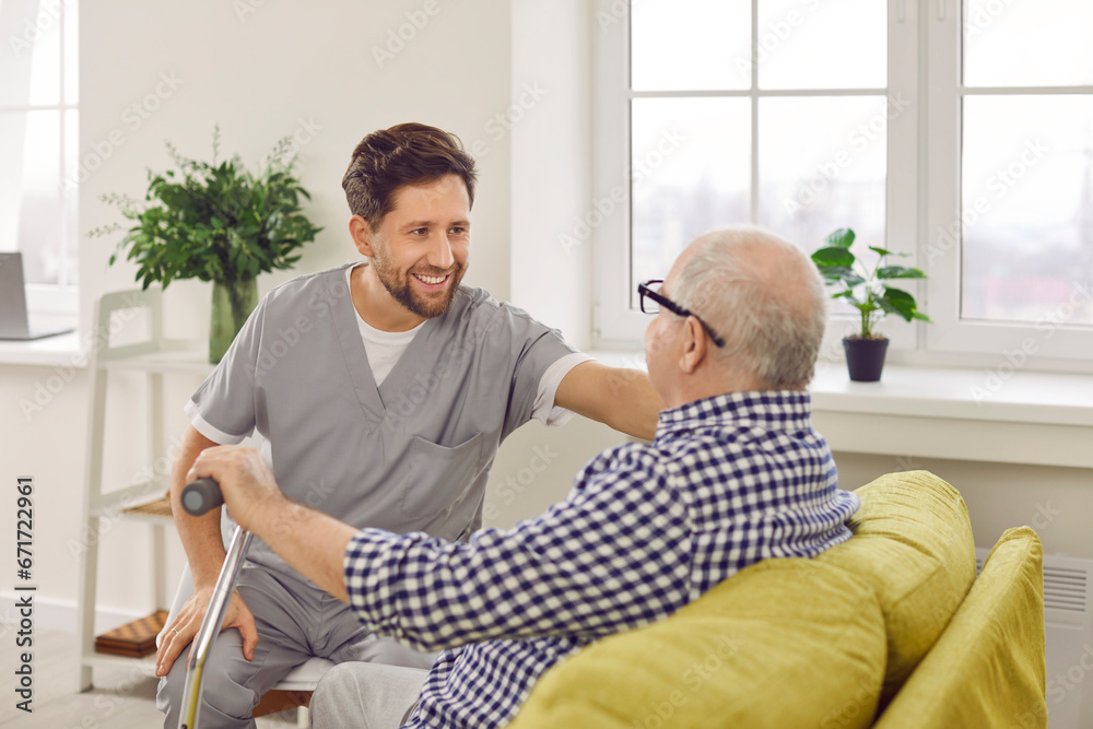 Optimistic male caregiver comforting senior patient, touching his shoulder. Male carer in uniform giving support to elderly man who sitting on sofa with walking cane. Elderly healthcare, medical care - obrazy, fototapety, plakaty 